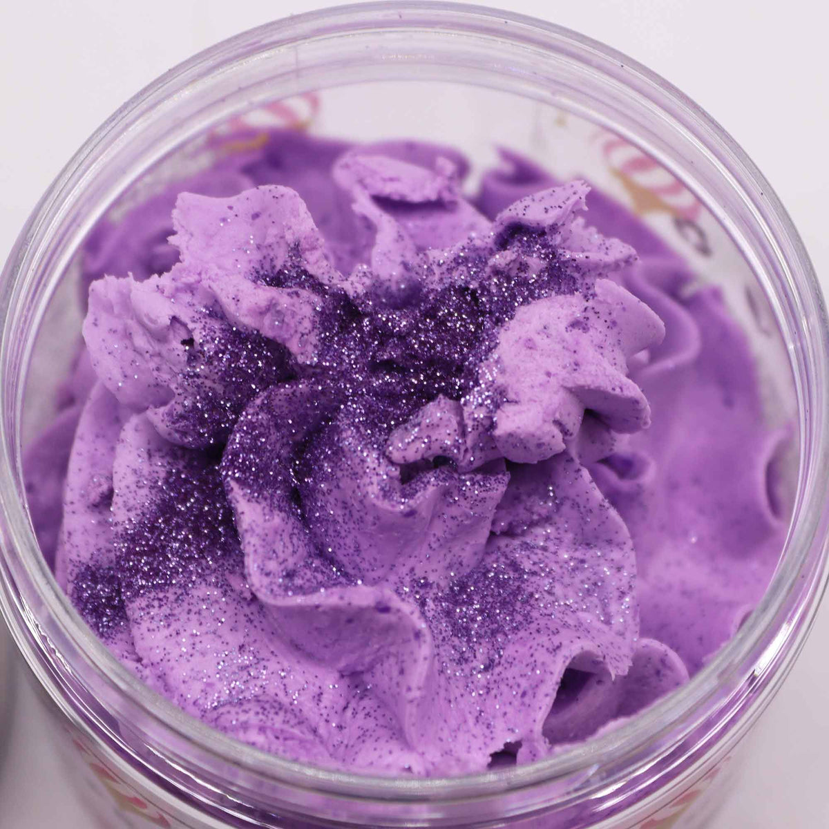 Purple whipped soap with glitter in a clear container on a white background
