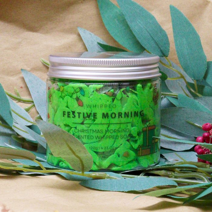 Green whipped soap in a jar with festive design on a beige background with green leaves.